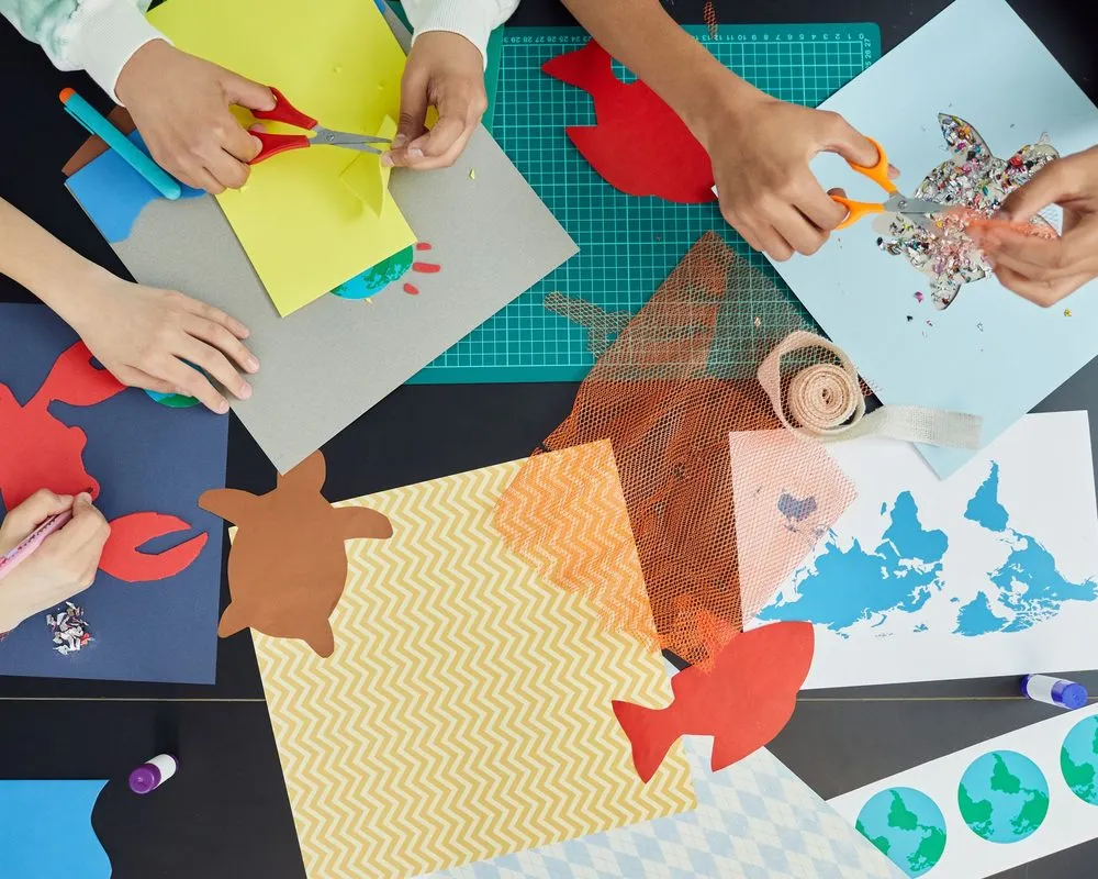 Hands cutting paper fish and turtles at a craft table covered in colored paper